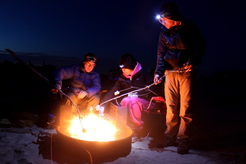 Three people gather around a campfire at night, roasting marshmallows with headlamps illuminating their faces.