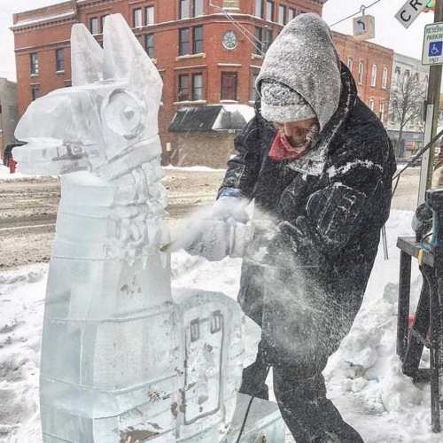 A person carves an ice sculpture of a llama in a snowy urban setting, surrounded by falling snowflakes.