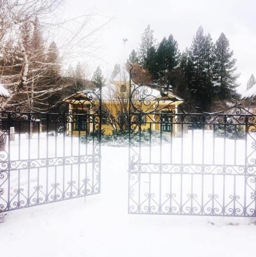 A snow-covered landscape with a yellow house behind ornate gates, surrounded by tall evergreen trees.