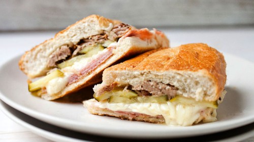 A close-up of two halves of a toasted sandwich filled with meat, cheese, and pickles on a white plate.