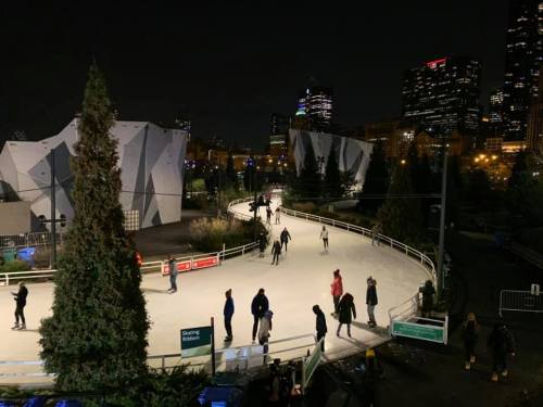 A nighttime ice skating rink with people skating, surrounded by trees and city buildings in the background.