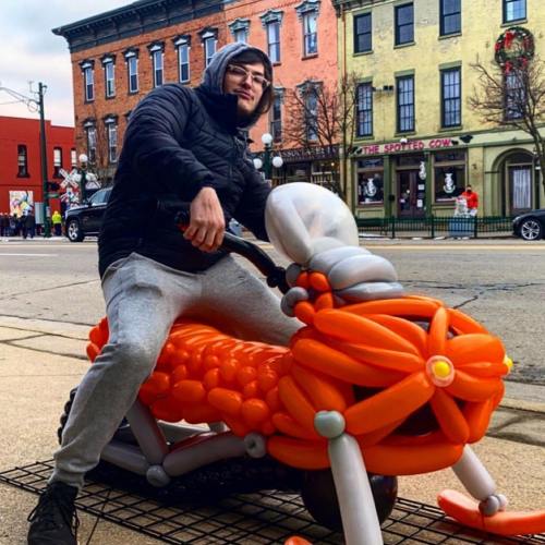 A person in a hoodie sits on a large orange balloon motorcycle on a city street.