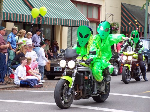 Three people in green alien costumes ride motorcycles in a parade, while onlookers watch from the sidewalk.