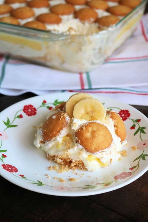A slice of banana pudding topped with vanilla wafers and banana slices on a floral plate, with a glass dish in the background.