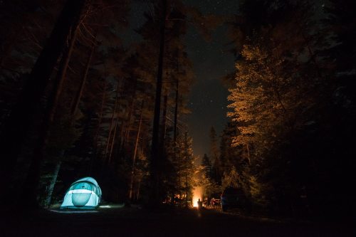 A glowing tent in a dark forest, with a campfire and trees illuminated under a starry night sky.