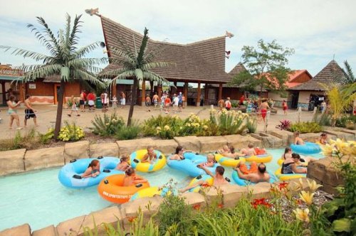 A tropical-themed water park entrance with guests floating on colorful tubes in a lazy river surrounded by lush greenery.