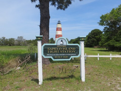 Sign for the historic Sapelo Island Light Station, with a lighthouse visible in the background and greenery surrounding it.