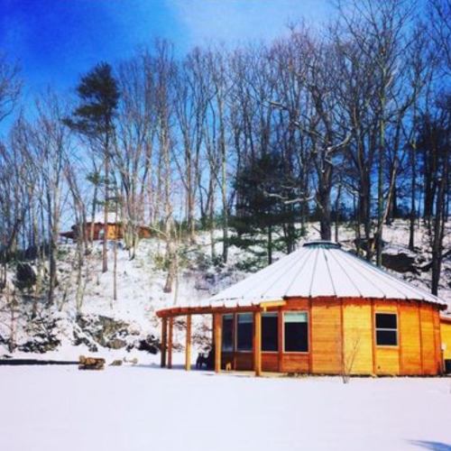 A wooden cabin with a circular design surrounded by snow and trees under a clear blue sky.