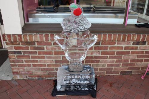 An ice sculpture of a large ice cream sundae with a cherry on top, displayed outside a shop.