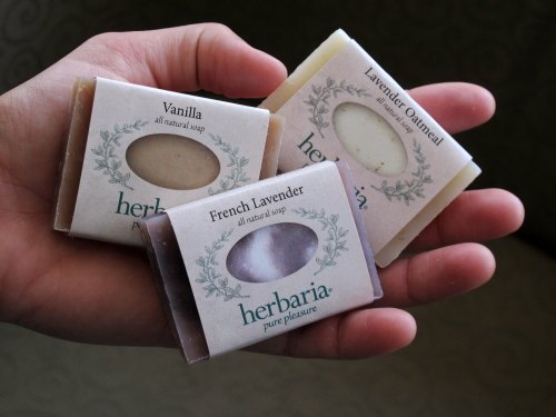 A hand holding three bars of all-natural soap: Vanilla, French Lavender, and Lavender Oatmeal, all from Herbaria.