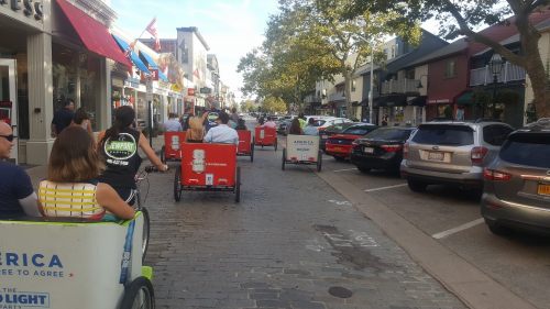A row of pedicabs on a busy street, with people enjoying a ride past shops and trees on a sunny day.