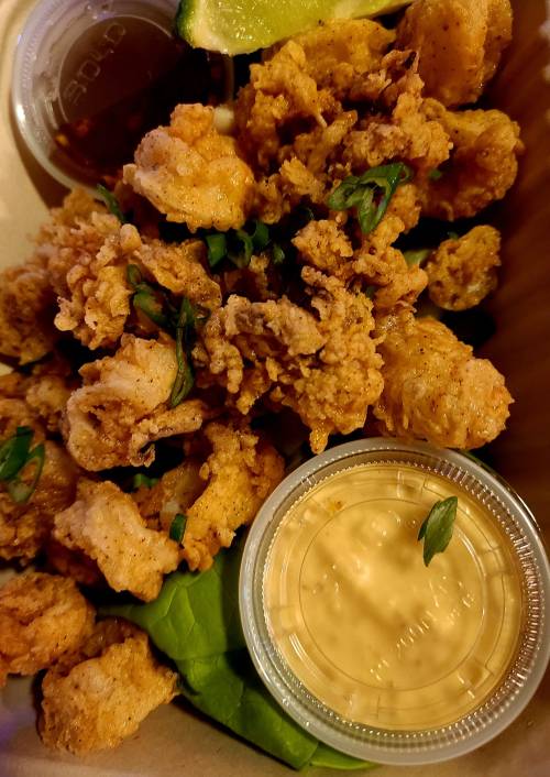 A close-up of crispy fried shrimp with dipping sauces and a lime wedge, served in a takeout container.