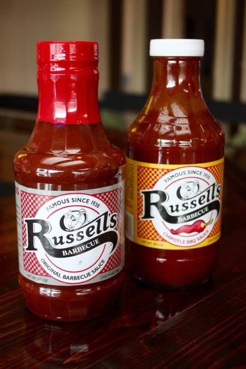 Two bottles of Russell's Barbecue Sauce: one original and one chipotle, both with distinctive labels on a wooden surface.