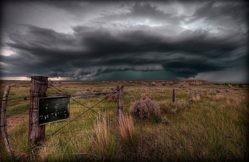A dramatic sky filled with dark storm clouds looms over a grassy landscape, with a rustic fence and sign in the foreground.