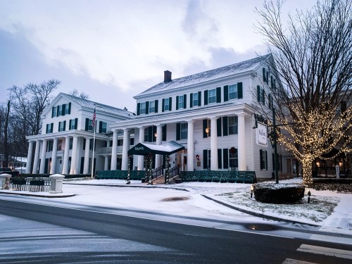 A historic white hotel with green shutters, surrounded by snow and holiday lights, under a cloudy winter sky.