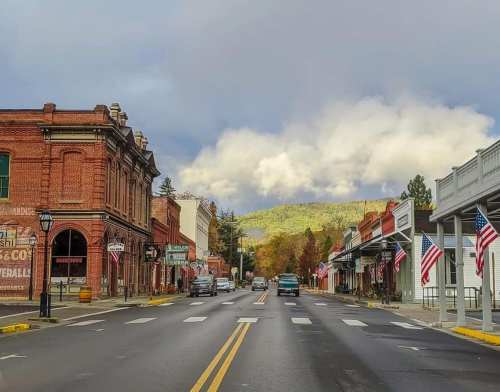 A quaint town street lined with brick buildings and American flags, set against a backdrop of mountains and cloudy skies.
