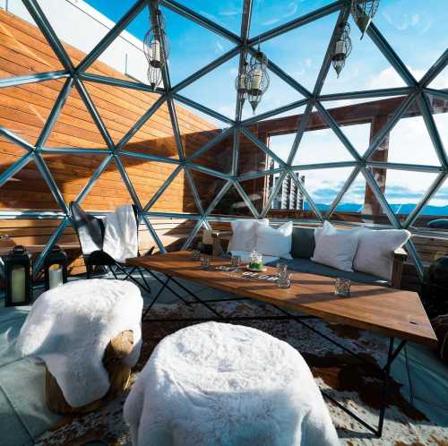 A cozy rooftop lounge with a glass dome, featuring a wooden table, plush seating, and scenic views.