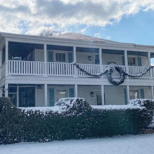 A white house with a porch, surrounded by snow and greenery, under a cloudy sky.