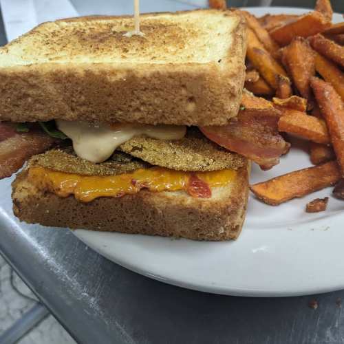 A sandwich with layers of bacon, cheese, and sauce, served with a side of sweet potato fries on a plate.