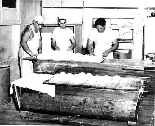 Three bakers in a vintage kitchen, working with dough on wooden tables. Flour and bread loaves are visible.