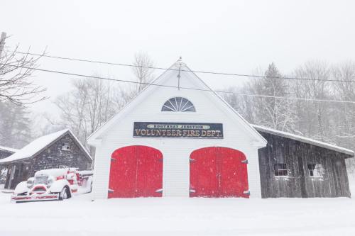 A snowy scene featuring a white fire station with red doors and a vintage fire truck in front.