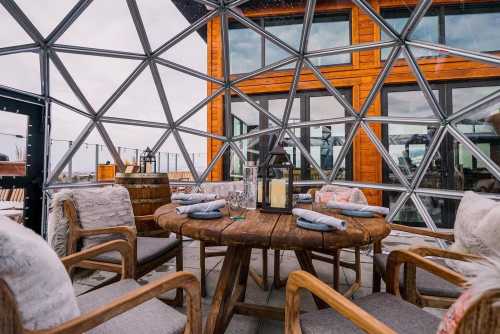 A cozy dining setup inside a geodesic dome, featuring a wooden table and chairs with soft blankets, overlooking a scenic view.