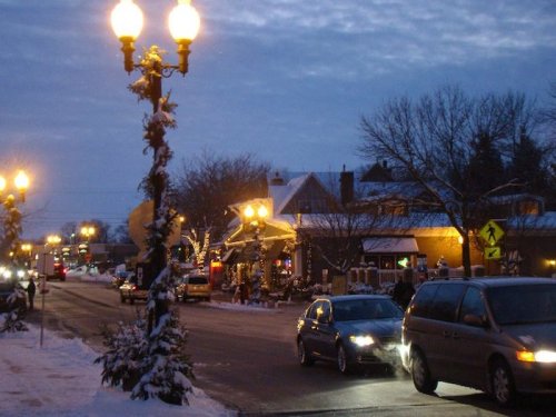 A snowy street scene at dusk, with cars, streetlights, and festive decorations on nearby buildings.
