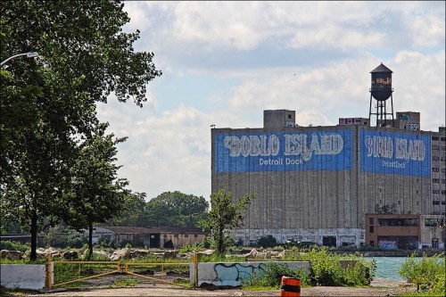 Abandoned industrial building with "GOBLO ISLAND" painted on the side, surrounded by overgrown vegetation and a water tower.