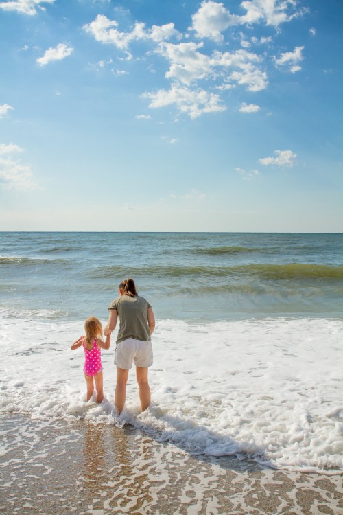 A woman and a young girl stand at the edge of the ocean, holding hands as waves gently wash over their feet.