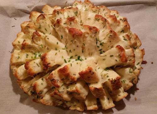 A round, baked bread topped with melted cheese and herbs, featuring a flower-like shape with cut sections.
