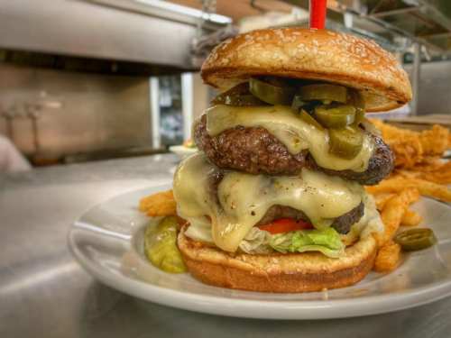 A towering burger with two patties, melted cheese, jalapeños, lettuce, and tomato, served with fries on a plate.
