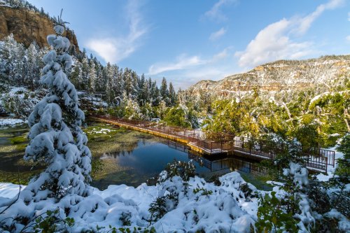 A snowy landscape with a wooden bridge over a calm pond, surrounded by trees and mountains under a blue sky.