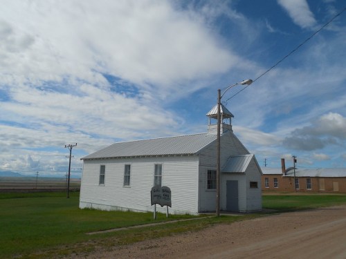A small white building with a peaked roof and bell tower, set against a cloudy sky and grassy landscape.