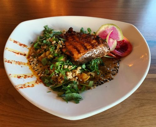 Grilled salmon atop a bed of greens, garnished with nuts, lime, and colorful radish slices on a white plate.
