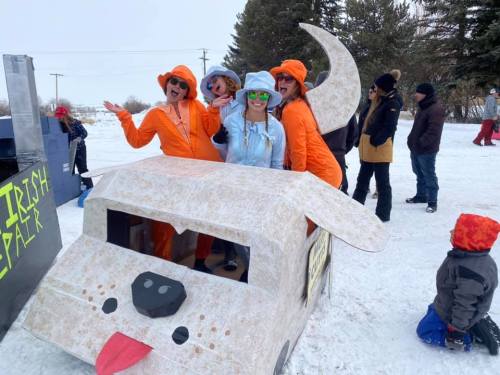 Five people in colorful outfits pose with a creatively decorated snow vehicle, surrounded by a snowy landscape.