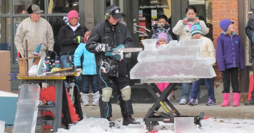 A man carves ice with a chainsaw while a crowd watches, bundled up in winter clothing. Snow is on the ground.