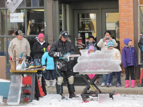 A man carves ice with a chainsaw while a crowd watches, bundled in winter clothing, in a snowy outdoor setting.