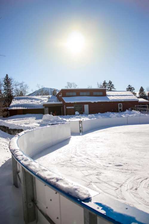 A sunny winter scene featuring a snow-covered ice rink and a red building in the background.