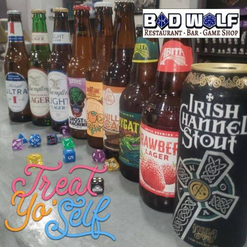 A variety of beer bottles lined up on a table with colorful dice and the text "Treat Yo' Self" in a playful font.