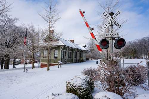 A snow-covered scene featuring a vintage train station, crossing signals, and bare trees under a cloudy sky.