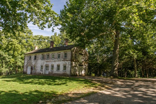 A weathered two-story house surrounded by large trees and green grass under a clear blue sky.