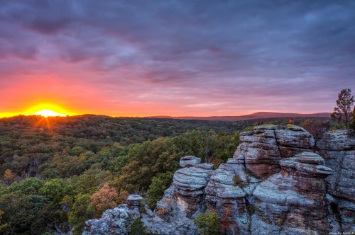 A vibrant sunset over a lush valley, with rocky formations in the foreground and colorful clouds in the sky.