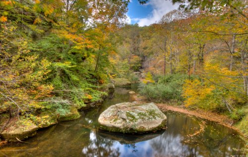 A serene river flows through a forest with vibrant autumn foliage and a large rock in the water.