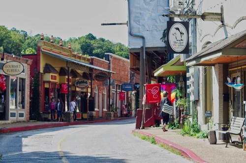 Charming street scene with colorful shops, flags, and people strolling in a quaint town. Lush greenery in the background.