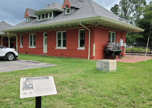 Historic building with red brick exterior, surrounded by grass, featuring a plaque and a vintage cart nearby.