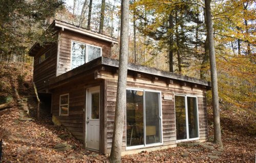 A modern wooden cabin nestled among autumn trees, featuring large glass windows and a sloped roof.