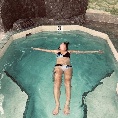 A person relaxes in a pool, floating on their back with arms outstretched, surrounded by stone walls.