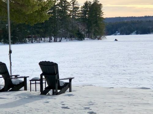Two black Adirondack chairs on a snowy lakeshore, with a snowmobile in the distance and trees lining the background.