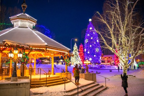 A festive winter scene with a gazebo, colorful Christmas lights, and snow-covered trees at night.