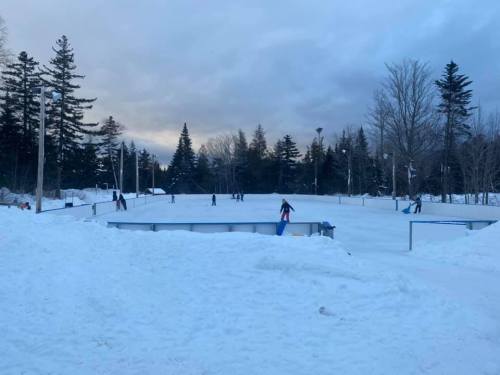 A snowy outdoor ice skating rink with people skating and trees in the background under a cloudy sky.